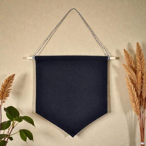 Könnte beinhalten: Ein dunkelblaues Filzbanner mit einem Holzstab und einer Schnur zum Aufhängen. Das Banner hat eine dreieckige Form am unteren Ende. Der Hintergrund ist ein neutrales Beige, mit getrockneten Pflanzen auf beiden Seiten.