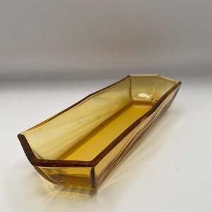 May include: A long, narrow, amber-coloured glass dish with a hexagonal shape and faceted sides. The vessel features a translucent, golden-yellow hue and a smooth, polished finish, with a flat base and angled, tapered ends that rise to a point.