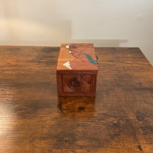 May include: A small, rectangular wooden box with a drawer and a decorative top. The box is stained a reddish-brown color. The top features a design with white, teal, and brown accents. A small, round knob is on the drawer.