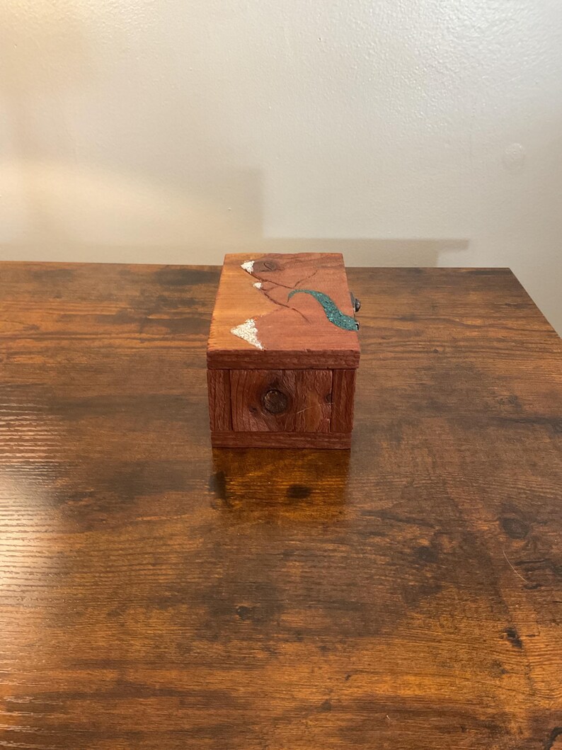 May include: A small, rectangular wooden box with a drawer and a decorative top. The box is stained a reddish-brown color. The top features a design with white, teal, and brown accents. A small, round knob is on the drawer.