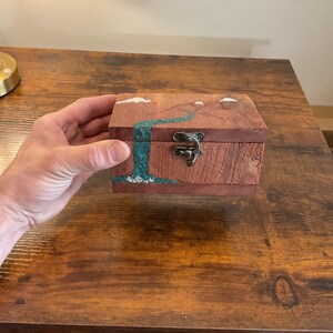 May include: A small, wooden box with a hinged lid and metal clasp. The box is made of reddish-brown wood and features a turquoise river design with white accents. The box is held by a person.