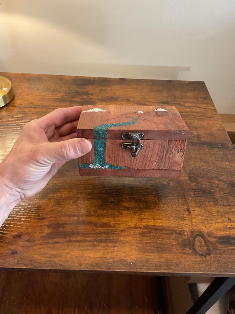 May include: A small, wooden box with a hinged lid and metal clasp. The box is made of reddish-brown wood and features a turquoise river design with white accents. The box is held by a person.