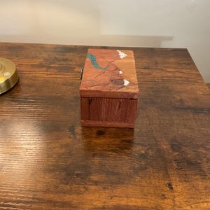 May include: A small, rectangular wooden box with a hinged lid. The box is made of reddish-brown wood with a natural grain. The lid features a turquoise and white decorative inlay. The box is sitting on a dark brown wooden surface.