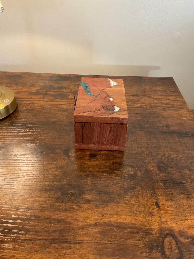 May include: A small, rectangular wooden box with a hinged lid. The box is made of reddish-brown wood with a natural grain. The lid features a turquoise and white decorative inlay. The box is sitting on a dark brown wooden surface.