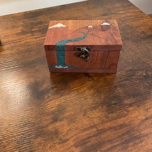 May include: A small, rectangular wooden box with a landscape painting of a waterfall and mountains. The box is made of reddish-brown wood and has a metal clasp. The waterfall is painted in shades of blue and green.