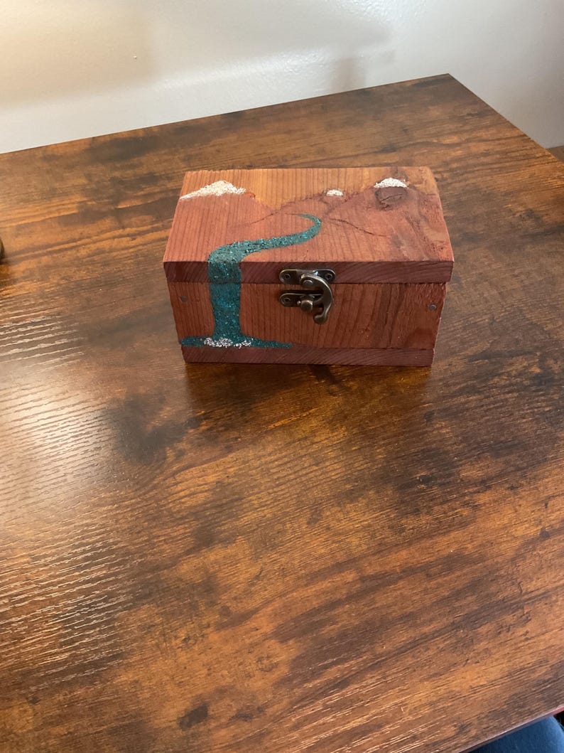 May include: A small, rectangular wooden box with a landscape painting of a waterfall and mountains. The box is made of reddish-brown wood and has a metal clasp. The waterfall is painted in shades of blue and green.
