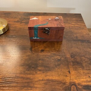 May include: A small, rectangular wooden box with a landscape design. The box is stained a reddish-brown color and features a teal waterfall and mountain scene. The box has a metal clasp and is sitting on a wooden surface.