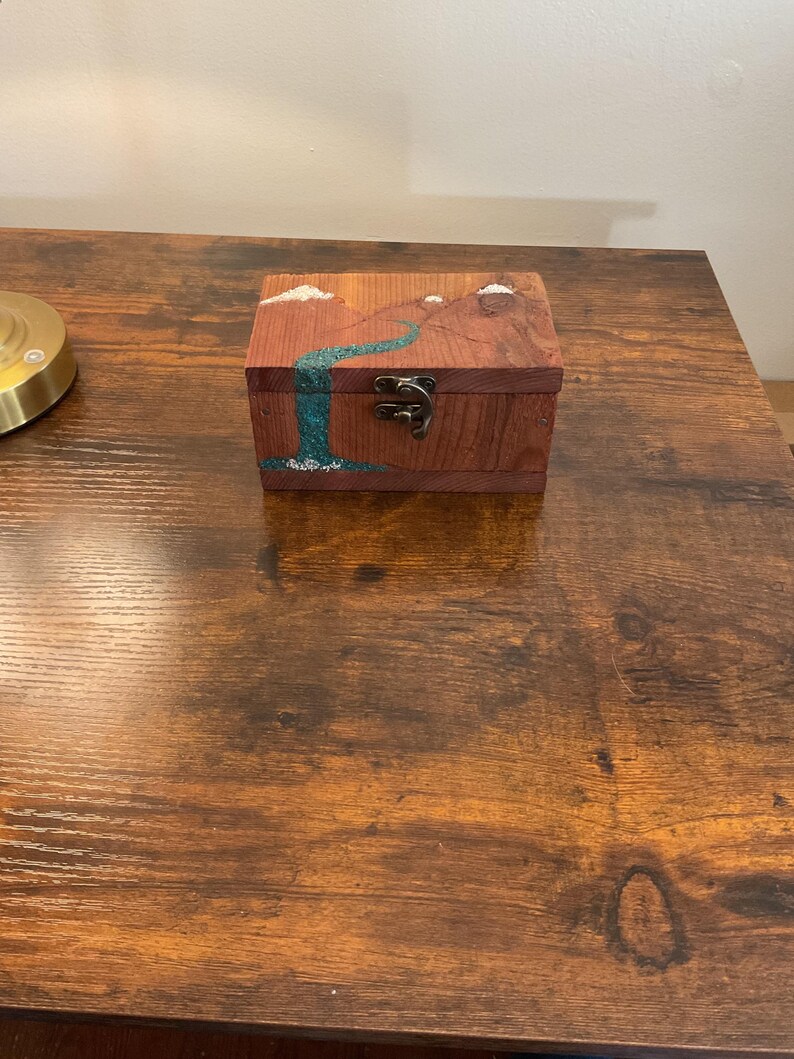 May include: A small, rectangular wooden box with a landscape design. The box is stained a reddish-brown color and features a teal waterfall and mountain scene. The box has a metal clasp and is sitting on a wooden surface.