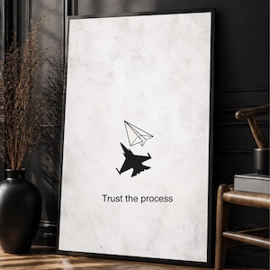 May include: A rectangular framed wall art piece with a white textured background. It features a black line drawing of a paper airplane above a solid black silhouette of a fighter jet. The text "Trust the process" is printed in black below the images.