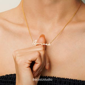 May include: A gold necklace featuring the name "Aurora" in a cursive script. The necklace is held by a person. The chain is fine, and the pendant is centrally placed. The background is a blurred view of a person's upper body.