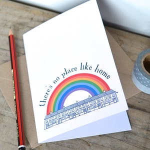 K&ouml;nnte beinhalten: Eine wei&szlig;e Karte mit einem Regenbogen und einer Reihe von H&auml;usern, die darauf gedruckt sind. Der Text "there's no place like home" ist in Schwarz &uuml;ber dem Regenbogen gedruckt.