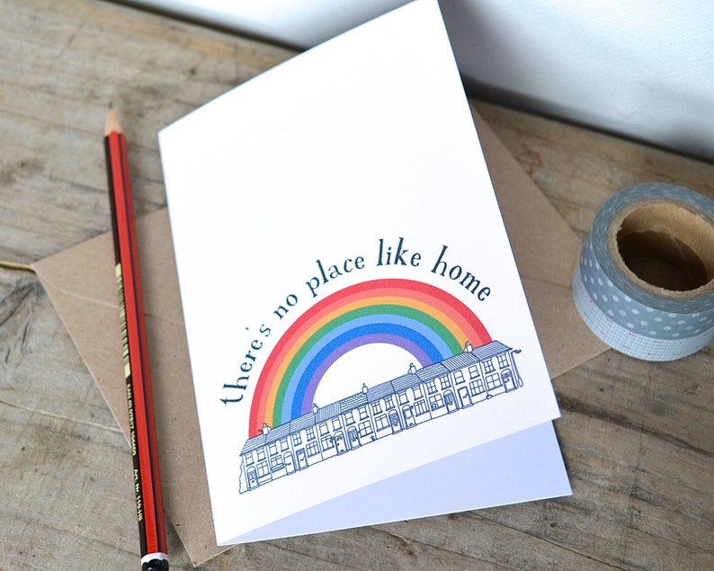 K&ouml;nnte beinhalten: Eine wei&szlig;e Karte mit einem Regenbogen und einer Reihe von H&auml;usern, die darauf gedruckt sind. Der Text "there's no place like home" ist in Schwarz &uuml;ber dem Regenbogen gedruckt.