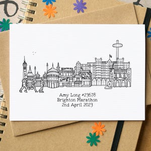 May include: A black and white line drawing of the Brighton skyline, featuring landmarks and runners. Text reads "Amy Long #23678 Brighton Marathon 2nd April 2023". The card is on a brown notebook.