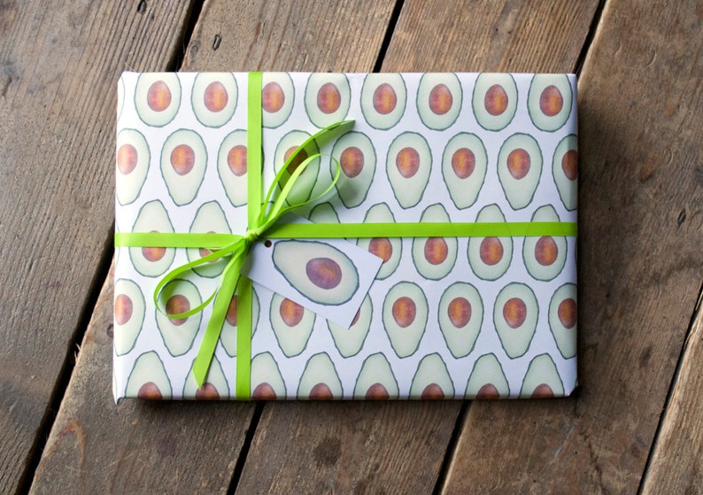 A rectangular gift, wrapped in white wrapping paper showing a repeat pattern of green avocados, cut in half. The gift has a lime green ribbon tied in a bow, and a small avocado gift tag.