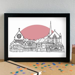 Könnte beinhalten: Schwarz gerahmter Kunstdruck mit einer detaillierten Schwarz-Weiß-Linienzeichnung einer Stadtlandschaft, einschließlich Gebäuden und einer Kirche, vor einem rosa kreisförmigen Hintergrund. Das Kunstwerk wird auf einer Holzoberfläche präsentiert.