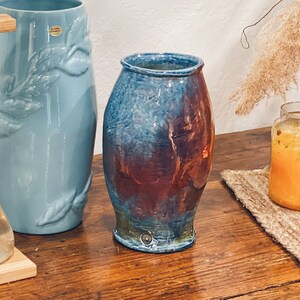 May include: A wooden stand with a clear glass vase holding white lilies, eucalyptus, and magnolia leaves.  A blue ceramic vase and a blue and brown ceramic vase are on a wooden table.