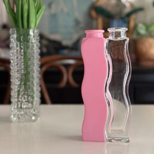 May include: A clear glass vase with a textured pattern holds pink tulips. Next to it are two decorative bottles, one pink and one clear, with a wavy design. The items are on a white surface.