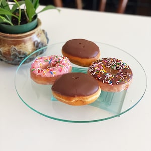 Puede incluir: Cuatro donuts en un soporte para pasteles de vidrio. Dos donuts glaseados con chocolate, un donut glaseado de color rosa con chispas de colores y un donut glaseado de color blanco con chispas de colores.