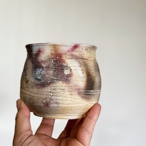 May include: A small, round, ceramic pot with a textured surface. The pot is decorated with a brown, white, and red glaze.