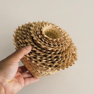May include: A woven basket with a spiral design. The basket is made of natural-colored straw and has a textured surface. The basket is held in a hand against a neutral background. The basket's design is reminiscent of a flower.