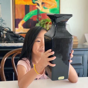 May include: A young person holds a black ceramic vase with a square shape and a label on the bottom. The vase is in front of a painting of a woman in a green dress.