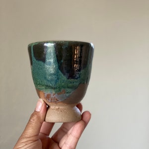 May include: A small, hand-thrown ceramic cup with a green, brown, and black glaze. The cup has a wide base and a rounded body.