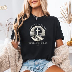 Puede incluir: Camiseta negra con un diseño gráfico circular que muestra una cruz sobre una colina, con el texto "FOR GOD SO LOVED THE WORLD THAT HE GAVE HIS ONLY SON JOHN 3:16". La camiseta es negra.