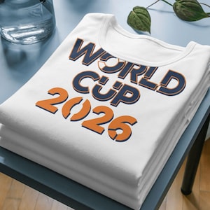 Folded white shirt displayed on a table featuring bold ‘World Cup 2026’ text design in blue and orange colors, shown as a mockup for soccer-themed apparel.