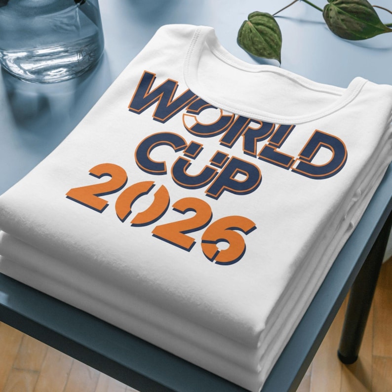 Folded white shirt displayed on a table featuring bold ‘World Cup 2026’ text design in blue and orange colors, shown as a mockup for soccer-themed apparel.