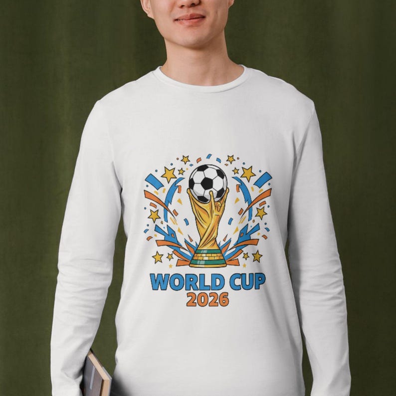 Person wearing a white long-sleeve shirt featuring a colorful soccer trophy illustration with a football on top, surrounded by stars and dynamic shapes, with ‘World Cup 2026’ text printed on the front.