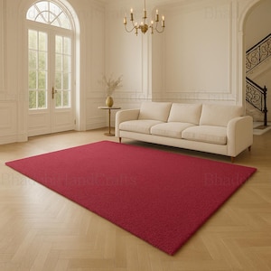 May include: A large, rectangular red rug dominates the foreground of a living room. The rug is a solid, deep red color and appears to be made of a soft, textured material. The room features a cream-colored sofa, a gold chandelier, and a large window.