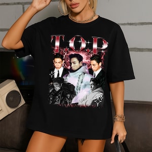 May include: Black t-shirt featuring a graphic design with the word "TOP" in a red and silver font. The design includes images of several people in suits and casual wear, with a red lightning bolt effect in the background.