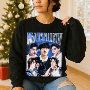 May include: Black sweatshirt featuring a graphic of five people with the name "MATTHEW" in a blue, stylized font. The image has a vintage aesthetic, with a blue background and the people in the image are wearing various outfits.