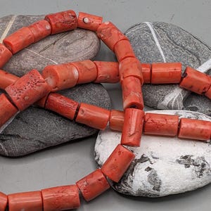 Op de afbeelding: Een ketting van koraalkralen. De kralen zijn fel oranjerood van kleur en cilindrisch van vorm. De ketting ligt op grijze stenen, tegen een lichtgrijze achtergrond. De kralen zijn ongeveer 1-2 cm lang.