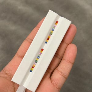 Puede incluir: Una tarjeta de presentación rectangular blanca con dos filas verticales de pequeñas piedras preciosas cuadradas en un degradado de arcoíris. Las piedras incluyen tonos de rojo, naranja, amarillo, verde, azul y morado en línea.
