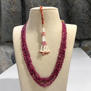 May include: A three-strand necklace featuring polished, oval-shaped deep pink beads. The necklace includes an adjustable red and gold cord closure finished with a decorative white and red beaded tassel hanging from the center of the strands.