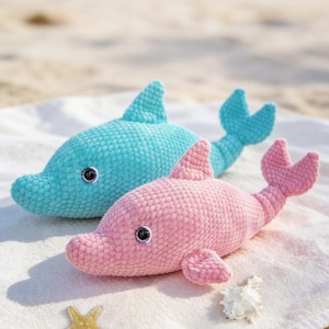 May include: Two crocheted dolphin plush toys, one turquoise and one pink, rest on a white towel on a sandy beach. Each dolphin has black eyes and a detailed fin and tail. A starfish and seashell are also on the towel.