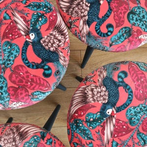 Puede incluir: Cuatro taburetes redondos con un vibrante estampado de pájaros y botánico. Los asientos están tapizados en tela coral con hojas turquesas y rosas y un pájaro azul estilizado. Cada taburete tiene patas negras cónicas.