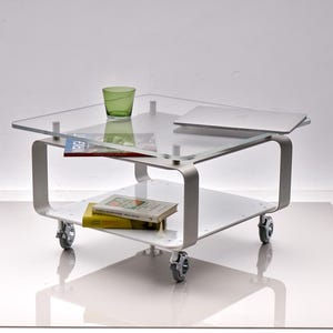 May include: A modern, rolling coffee table with a clear glass top and a lower shelf. The table has a white frame with curved metal supports and small wheels. A green glass and books are on the top shelf, and a laptop is also present.