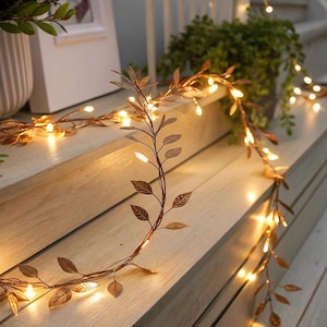 Könnte beinhalten: Eine dekorative Girlande mit bronzefarbenen Blättern und kleinen, warmweißen LED-Lichtern. Die Girlande ist über Holzstufen drapiert und schafft eine warme Atmosphäre. Die Blätter weisen detaillierte Adern auf.