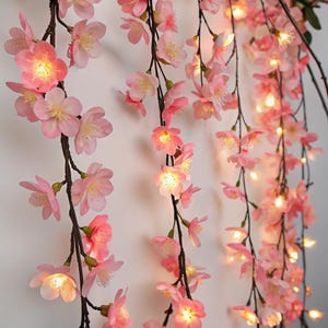 Puede incluir: Guirnalda de luces de flores de cerezo rosas. Las flores artificiales son de color rosa suave con centros amarillos, ensartadas en ramas marrones oscuras. Las luces son de color amarillo cálido, creando un brillo suave. Guirnalda floral decorativa.