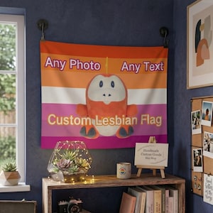 May include: A custom lesbian flag with the text "Any Photo" and "Any Text" in orange and white. A plush toy is centered on the flag. The flag is hanging on a wall above a wooden bookshelf with books, a camera, and a terrarium.