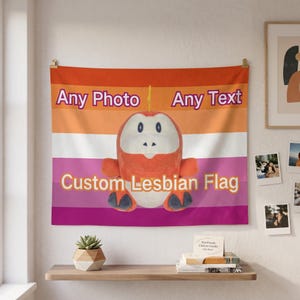 Puede incluir: Una bandera lésbica personalizada con rayas naranjas, blancas y rosas, con el texto "Any Photo Any Text" y "Custom Lesbian Flag". Un peluche con forma de criatura está centrado en la bandera.