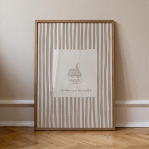 May include: Framed art print featuring a hand-drawn house illustration with the text "this house you'll love itself" on a striped background. The frame is made of light brown wood, and the artwork is displayed on a wooden floor.
