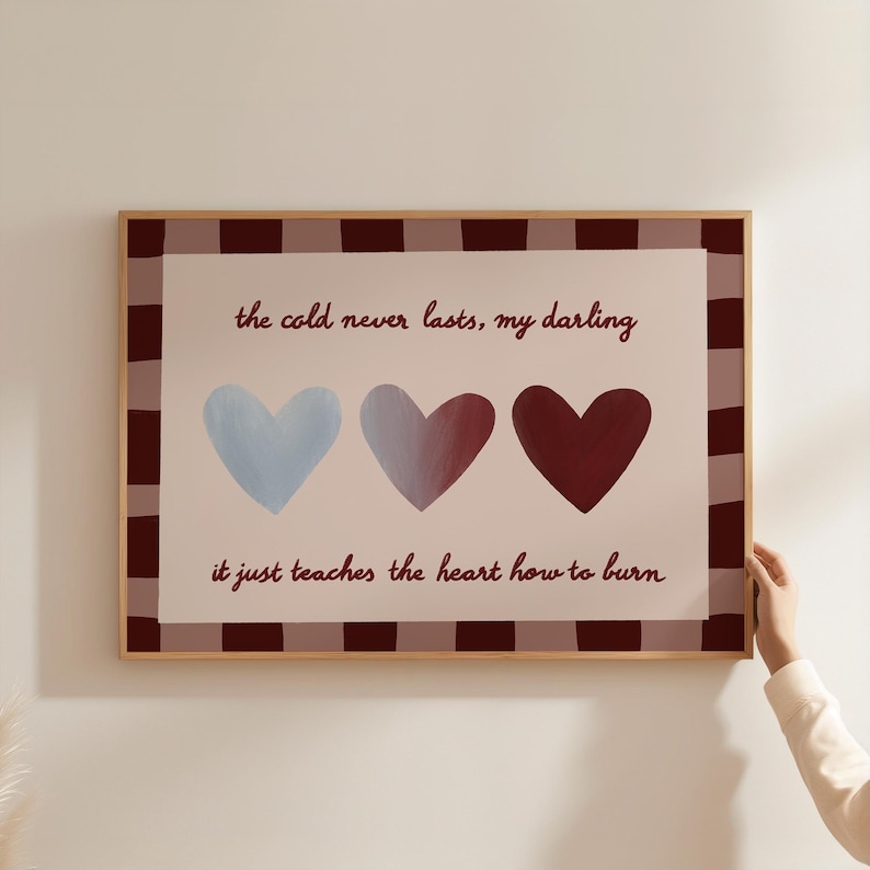 Puede incluir: Impresi&oacute;n enmarcada con el texto "the cold never lasts, my darling, it just teaches the heart how to burn." La obra presenta tres corazones en tonos azules, morados y rojos. El marco tiene un borde a cuadros marr&oacute;n y blanco.