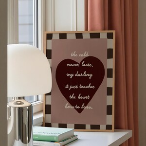 May include: A framed print with a light wood frame features the text "the cold never lasts, my darling, it just teaches the heart how to burn" over a burgundy heart. The background is a pinkish-brown color with a black and white checkered border.