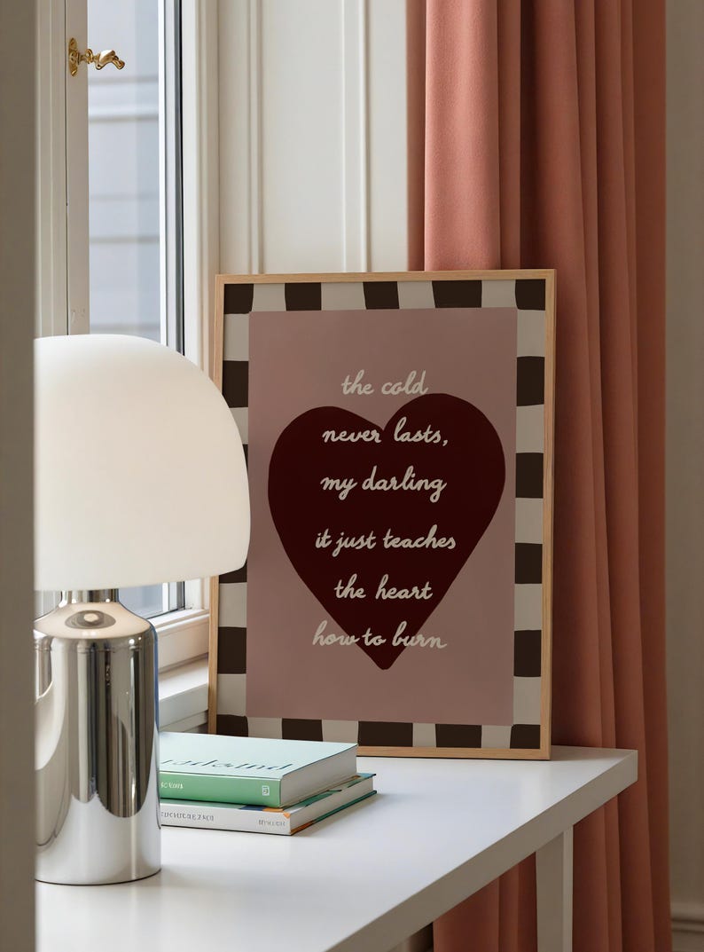 May include: A framed print with a light wood frame features the text "the cold never lasts, my darling, it just teaches the heart how to burn" over a burgundy heart. The background is a pinkish-brown color with a black and white checkered border.