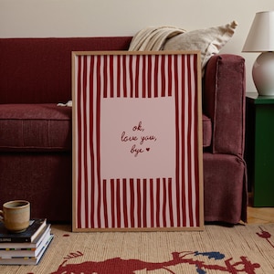 May include: A framed art print with a wooden frame. The print features a pink square with the words "ok, love you, bye" in black script. The background has vertical red stripes. The print is displayed on a patterned rug.