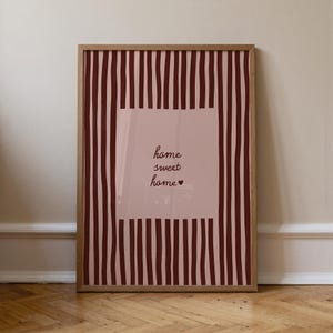 May include: A framed print with a wooden frame. The print features a pink rectangle with the words "home sweet home" in a handwritten font. The background has vertical burgundy stripes.