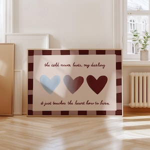 Puede incluir: Un estampado enmarcado con tres corazones en tonos azul, gris y burdeos. El estampado incluye el texto "el fr&iacute;o nunca dura, cari&ntilde;o, solo le ense&ntilde;a al coraz&oacute;n a arder". El marco tiene un borde a cuadros marr&oacute;n y blanco.